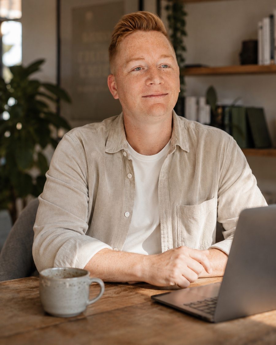Marco Engel am Holztisch mit Laptop und Kaffeetasse, lockeres Cordhemd